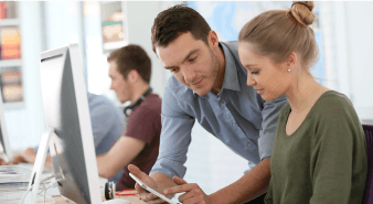 Female sat at computer as male teacher demonstrates tablet